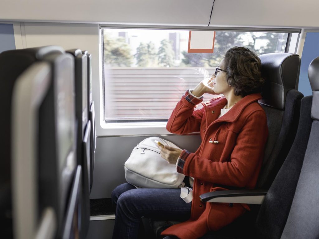 Smiling woman in red duffle coat with on smartphone sits near window in suburban train. Travel by land vehicle.